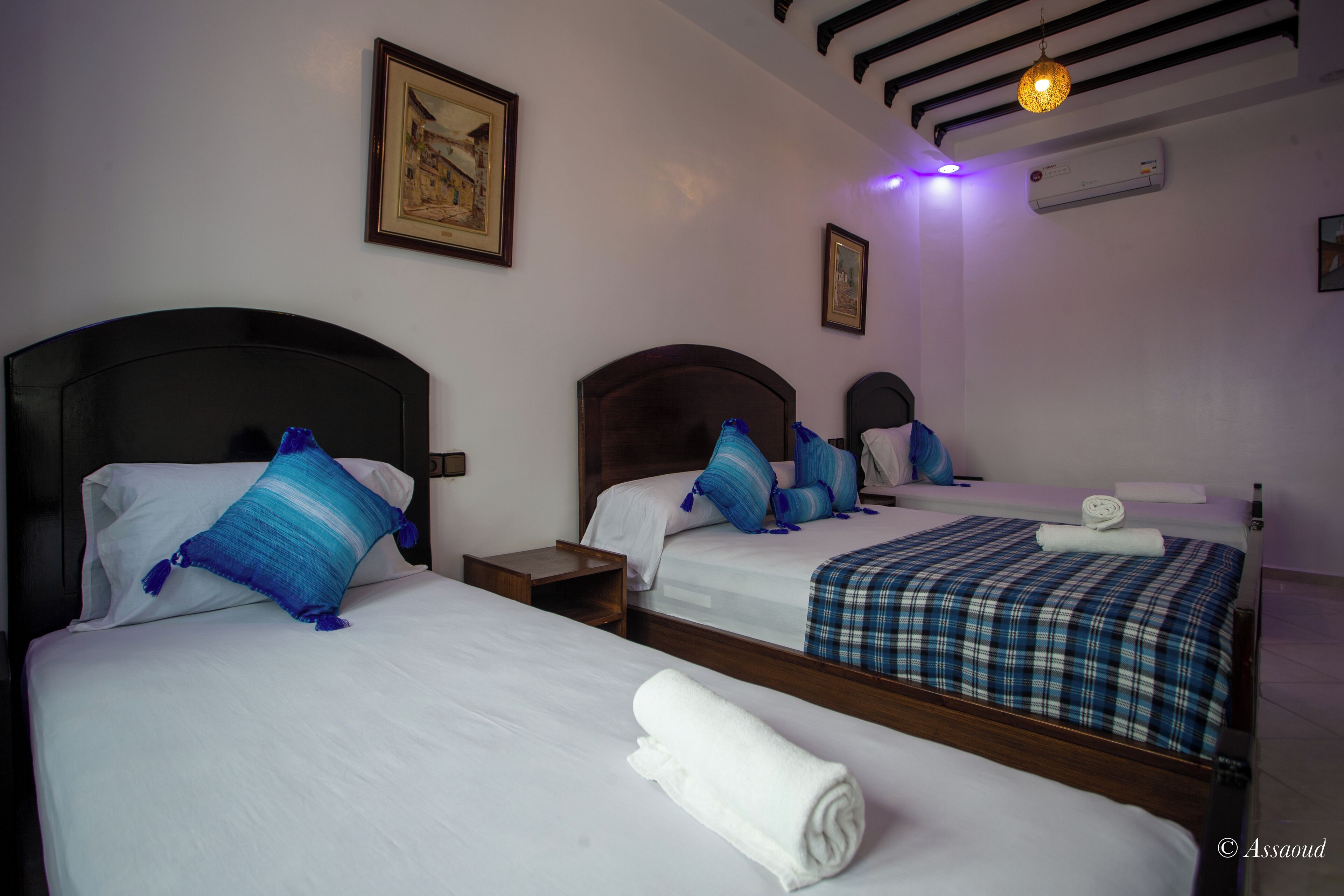 Ein Hotelzimmer mit zwei Betten, jeweils mit Kopfteil, Nachttisch und einem zusammengerollten Handtuch auf dem Bett. An der Wand hängt ein gerahmtes Bild und es befindet sich eine Klimaanlage im Raum.