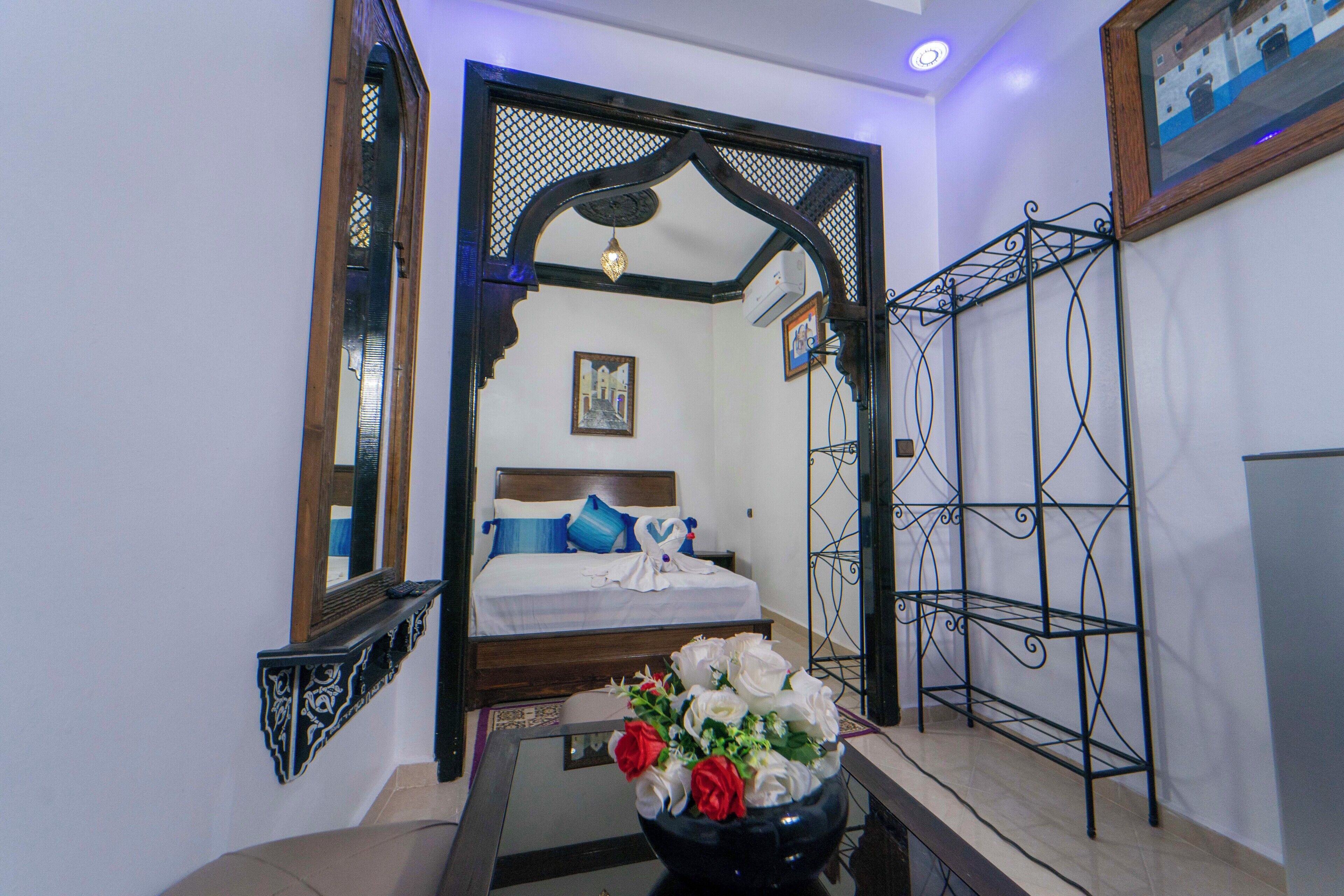 Un dormitorio con una cama, un espejo decorativo, una estantería de metal negro y un florero con flores.