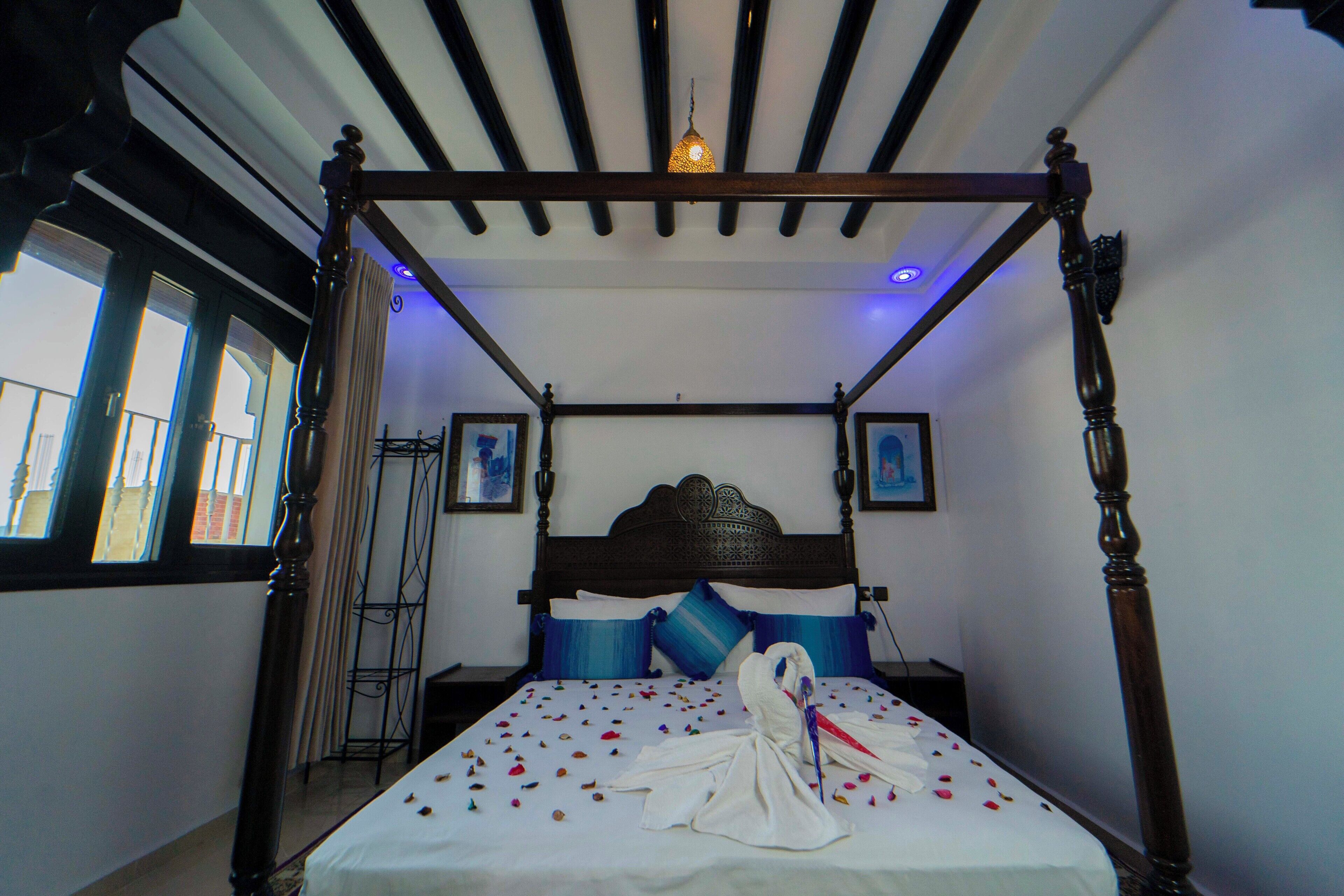 A four-poster bed with a white bedspread and blue pillows, a wooden headboard, and a hanging light fixture.