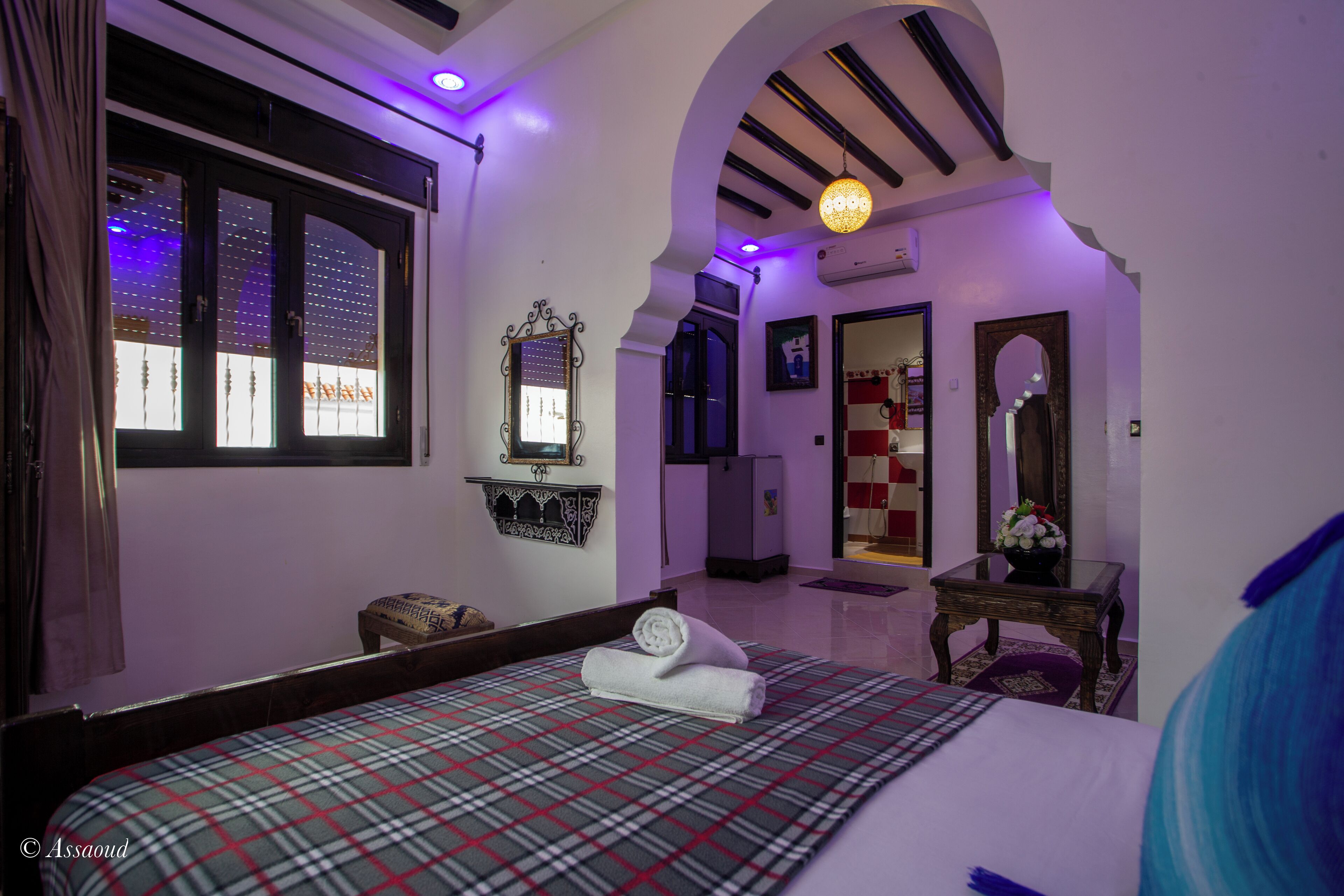 A bedroom with a plaid bedspread, a small stool, a mirror, and a window with curtains.