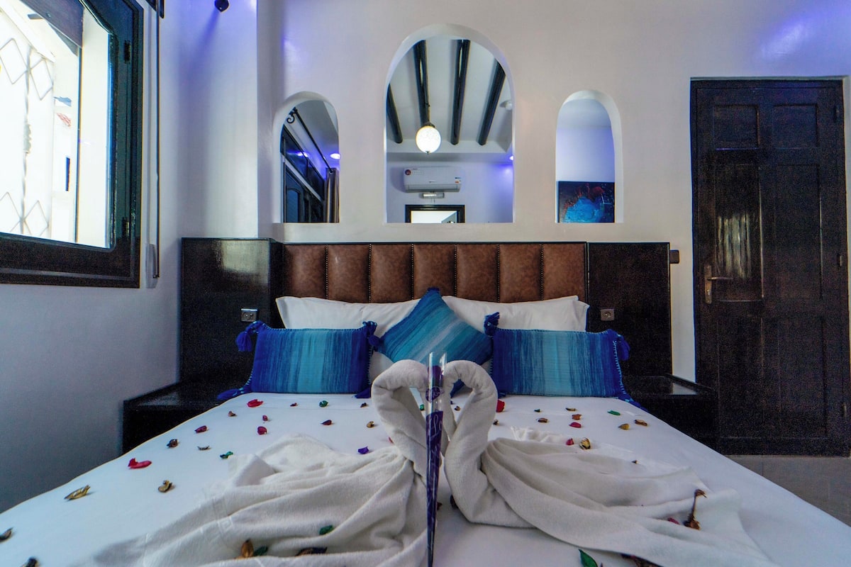 Una habitación de hotel con una cama grande, almohadas a rayas azules y blancas, un cabecero de madera y una ventana con vistas.