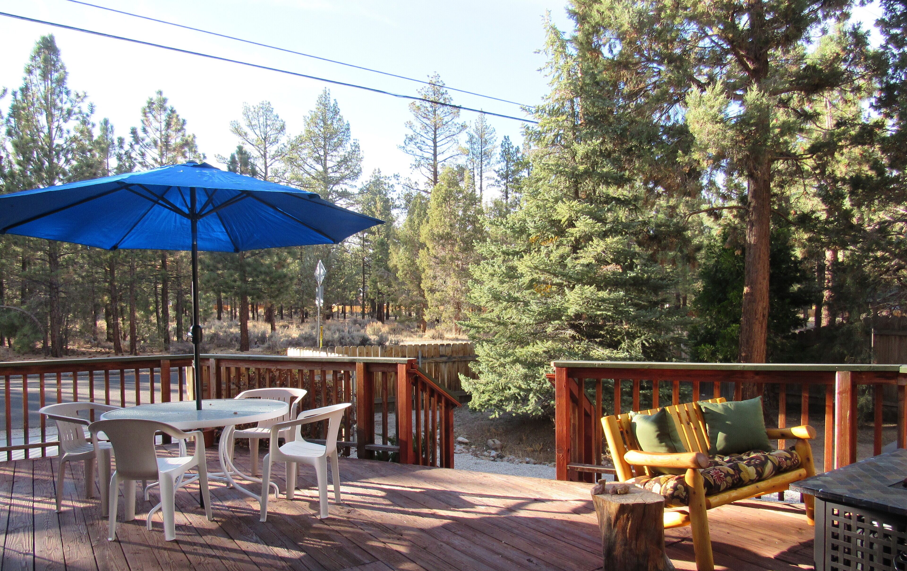 A wooden deck with a table and chairs, a blue umbrella, and a wooden bench with cushions.