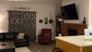 Ein Wohnzimmer mit einer schwarzen Ledersofa, einem roten Sessel, einem Holztisch und einem kleinen Hocker. Ein Flachbildfernseher ist an der Wand über einem Kamin aus Ziegeln angebracht.