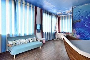 A room with a sofa, a small table, a chair, a large window with curtains, and a mural of an underwater scene.