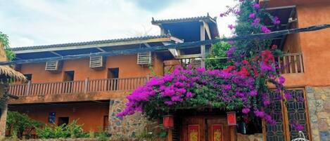 A traditional building with a wooden door and balcony, surrounded by lush greenery and vibrant flowers.