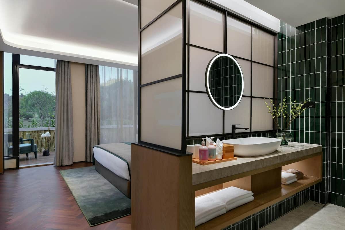 Ein modernes Hotelzimmer mit einem großen Bett, einem hölzernen Waschtisch mit rundem Spiegel und Blick ins Freie durch eine Schiebetür.