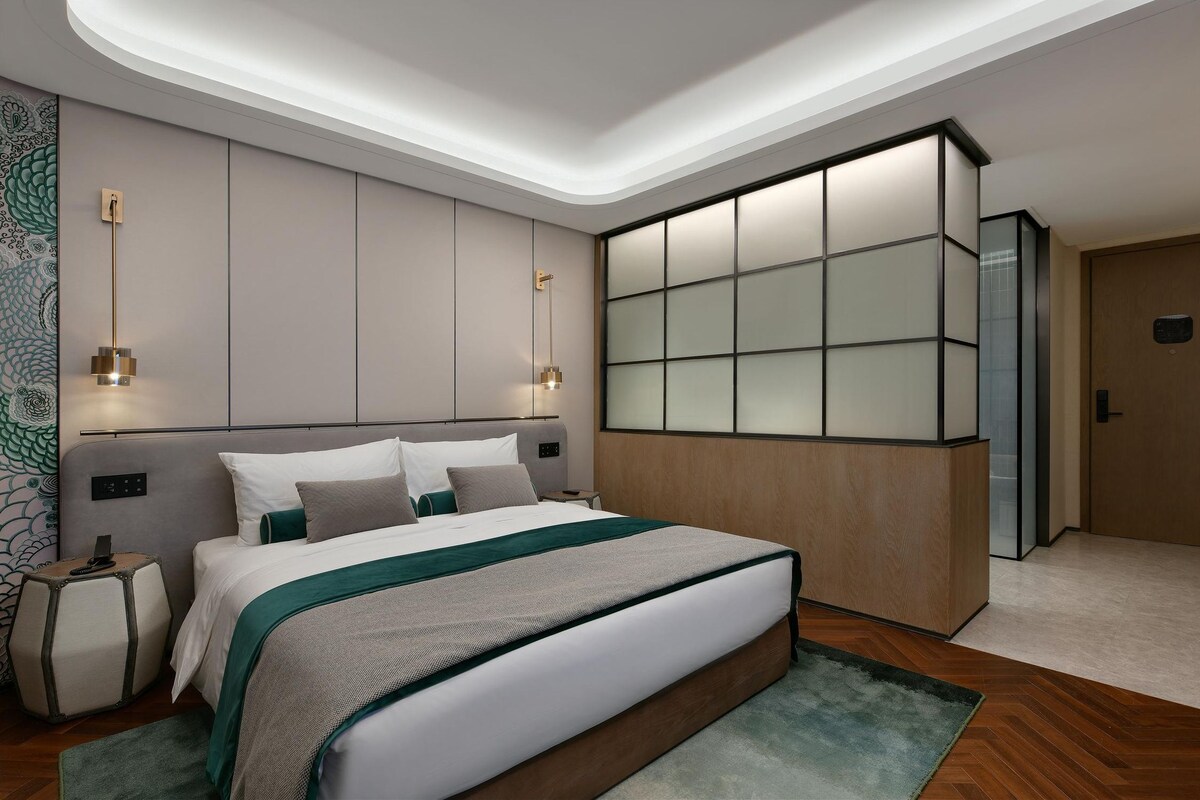 A modern bedroom with a large bed, a nightstand, a wall-mounted light, and a sliding glass door.