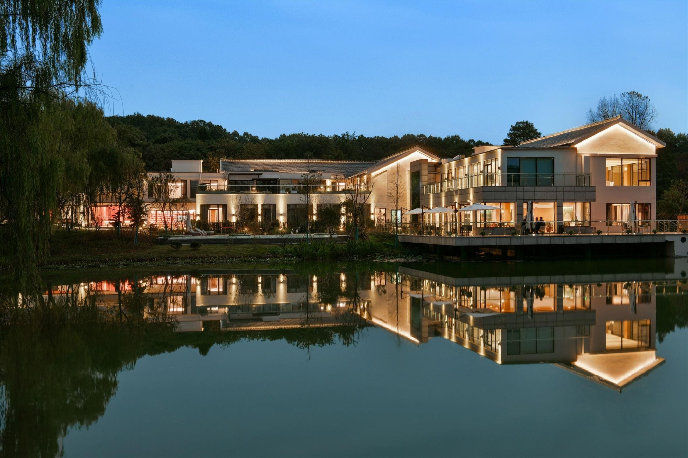 A modern hotel with a reflective pool, multiple buildings, and a forested hill in the background.