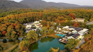 A residential area with modern houses, a central pond, and a backdrop of autumn-colored trees and hills.