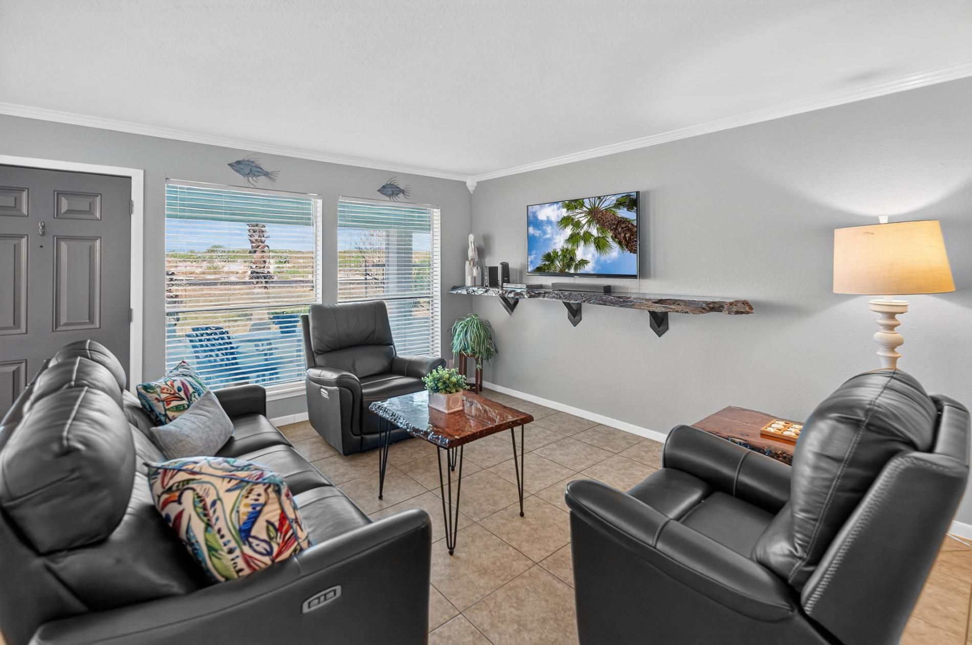 A living room with a sofa, armchairs, a coffee table, and a TV. There are windows with blinds, a door, and a wall-mounted shelf with decorative items.