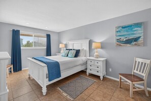 Une chambre à coucher avec un lit, des tables de chevet, une chaise, une fenêtre avec des rideaux, un tableau et un tapis.