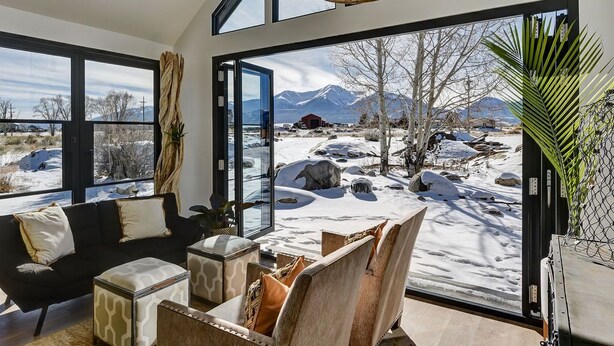 A modern living room with a large glass door leading to a snowy outdoor area, featuring a sofa, ottoman, and chairs. - Riverfront Retreat w/ Mountain Views (Buena Vista)