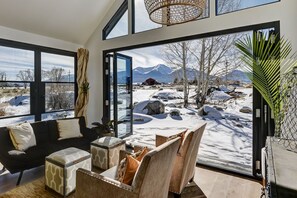 Un salon moderne doté d’une grande porte vitrée donnant sur un espace extérieur enneigé, et comprenant un canapé, un pouf et des fauteuils.