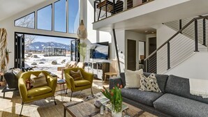 A spacious living room with a large window, a grey sofa, a yellow armchair, and a coffee table with decorative items. - Riverfront Retreat w/ Mountain Views (Buena Vista)