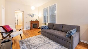 A living room with a gray sofa, a white armchair, a fireplace, and a large window with blinds. - Cozy two bedroom close to Wrigley Field (Chicago)