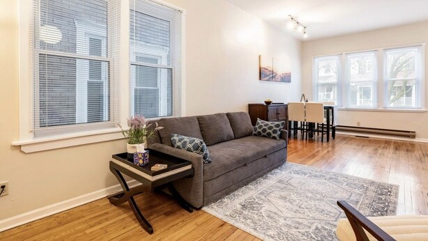 A living room with a grey sofa, a small side table with a plant, and a dining area with a table and chairs. - Cozy two bedroom close to Wrigley Field (Chicago)