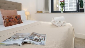 A neatly made bed with a magazine, a window with a view, and a plant on the windowsill.