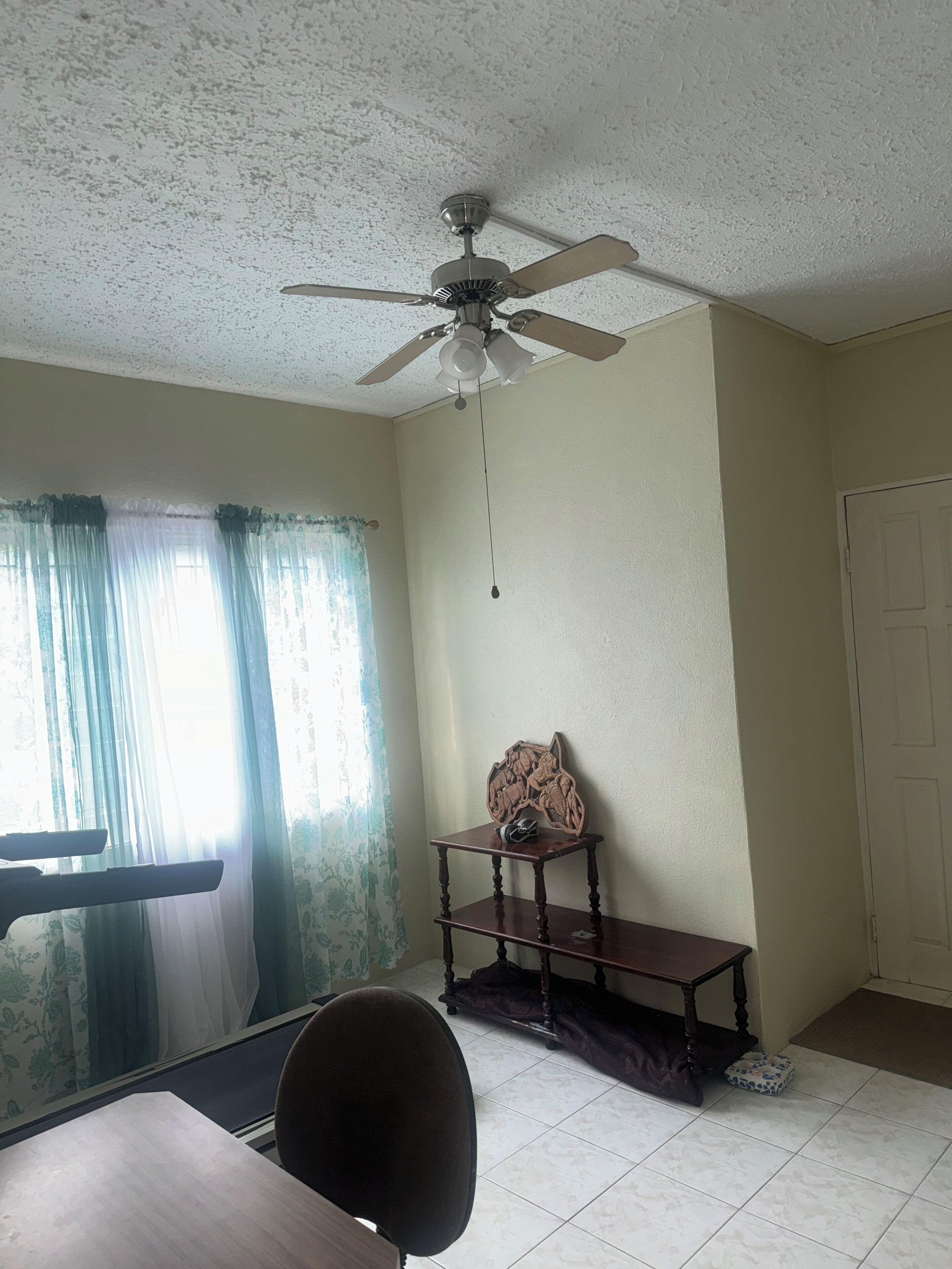 A room with a ceiling fan, a table, a chair, a decorative wall piece, and a door.