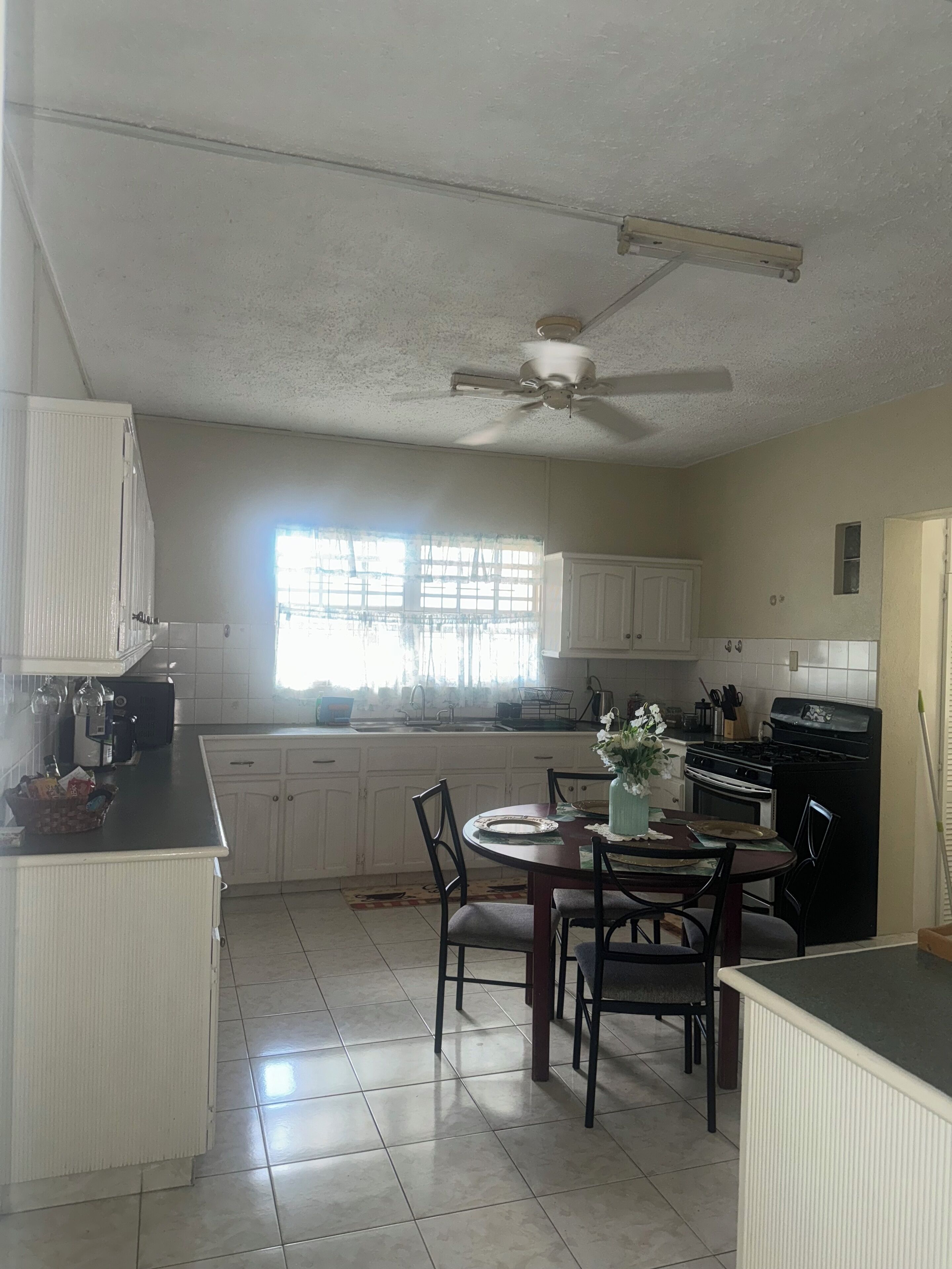 A kitchen with white cabinets, a dining table with chairs, a ceiling fan, and a window with curtains.