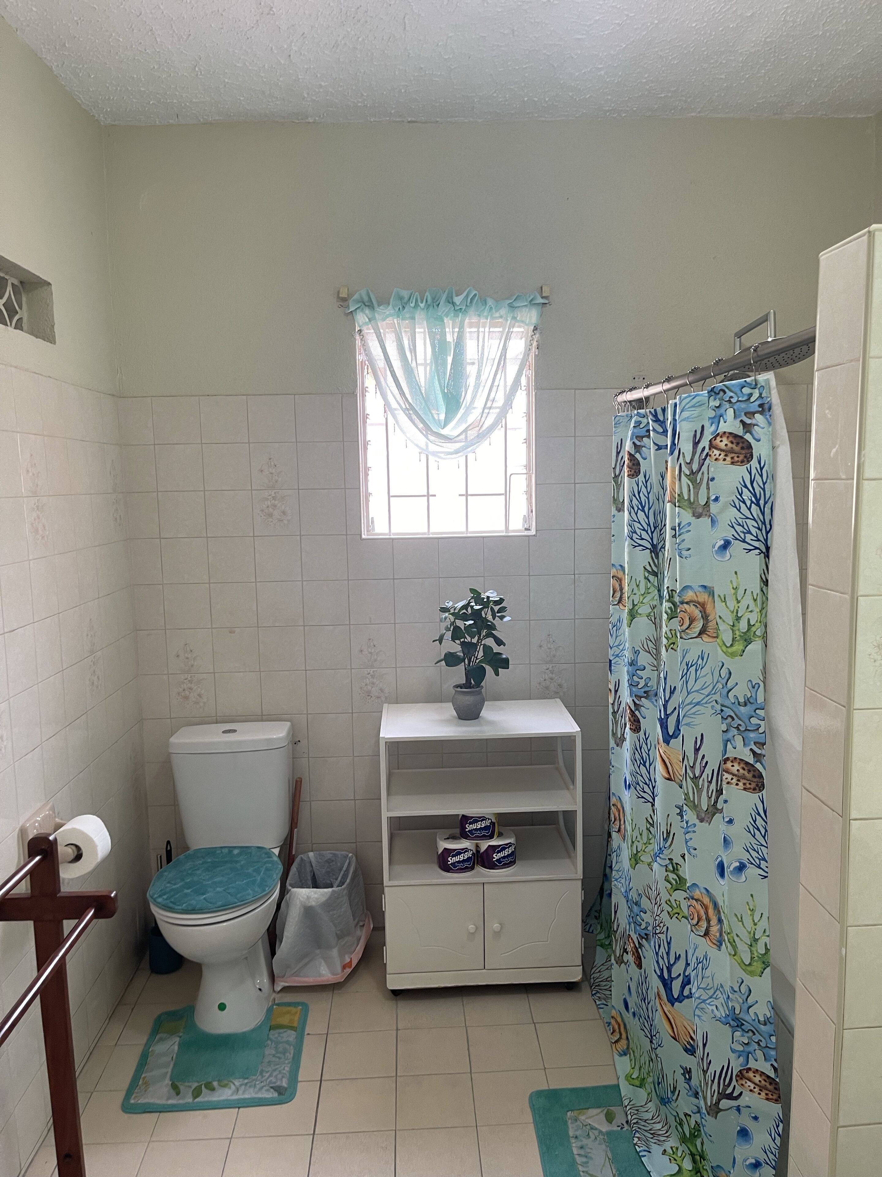 A small bathroom with a toilet, a shelf with a potted plant, a shower curtain with a marine life pattern, and a window with a sheer curtain.