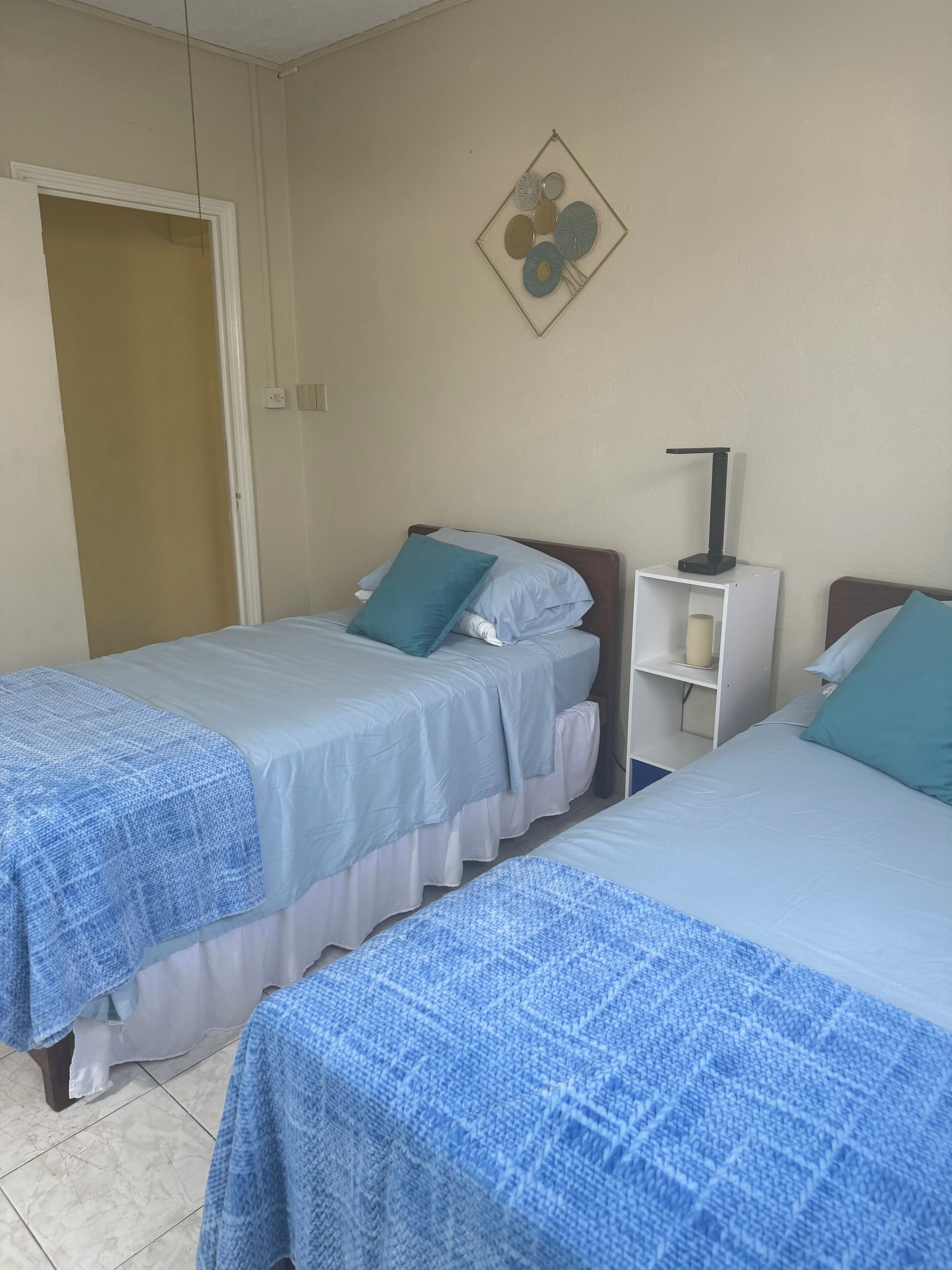 A hotel room with two beds, each with a blue bedspread and pillows, a small white nightstand with a lamp, and a wall decoration above the beds.
