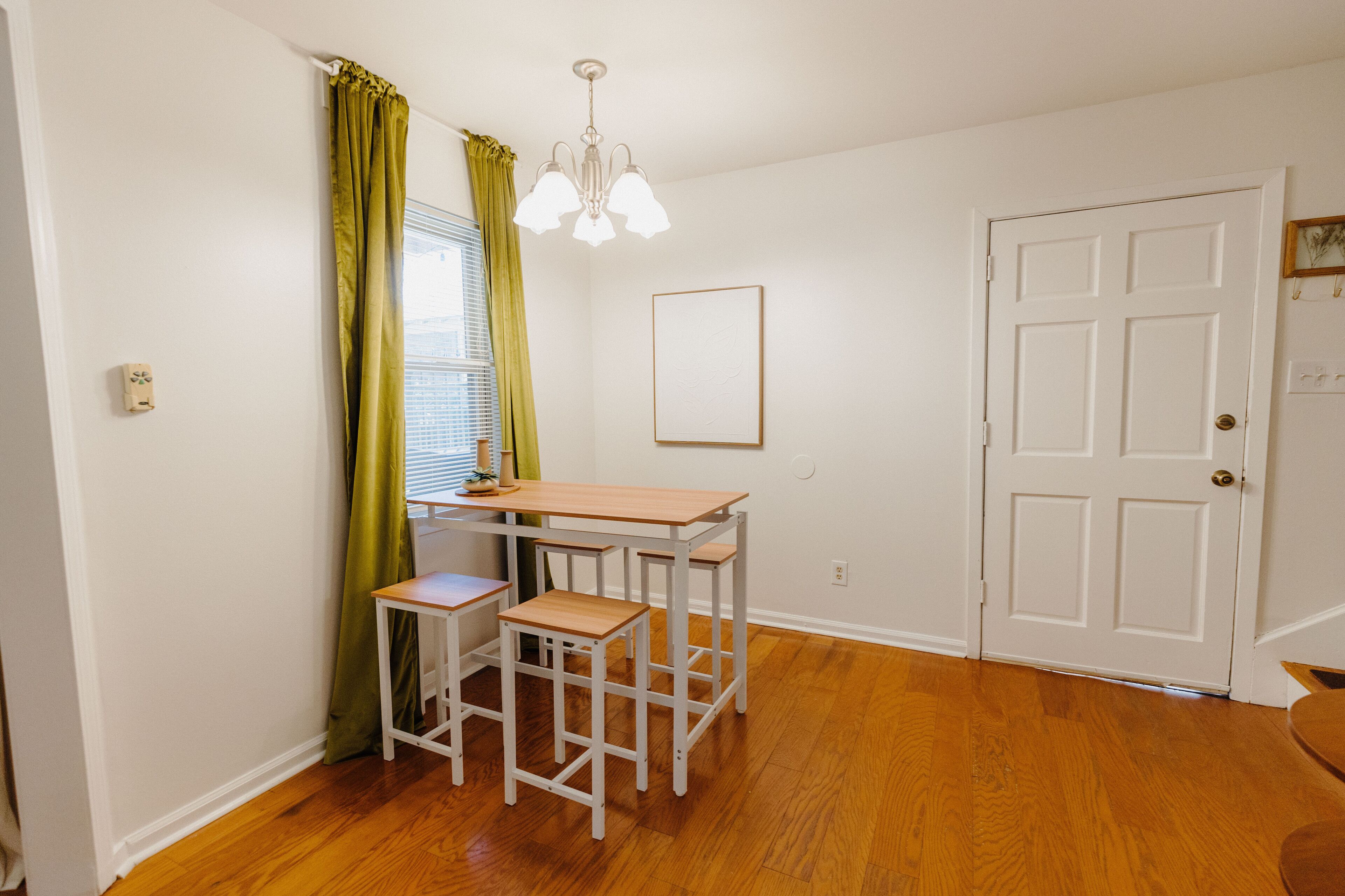 Une pièce avec un plancher en bois, une porte blanche, une table avec deux chaises et une fenêtre ornée de rideaux verts.