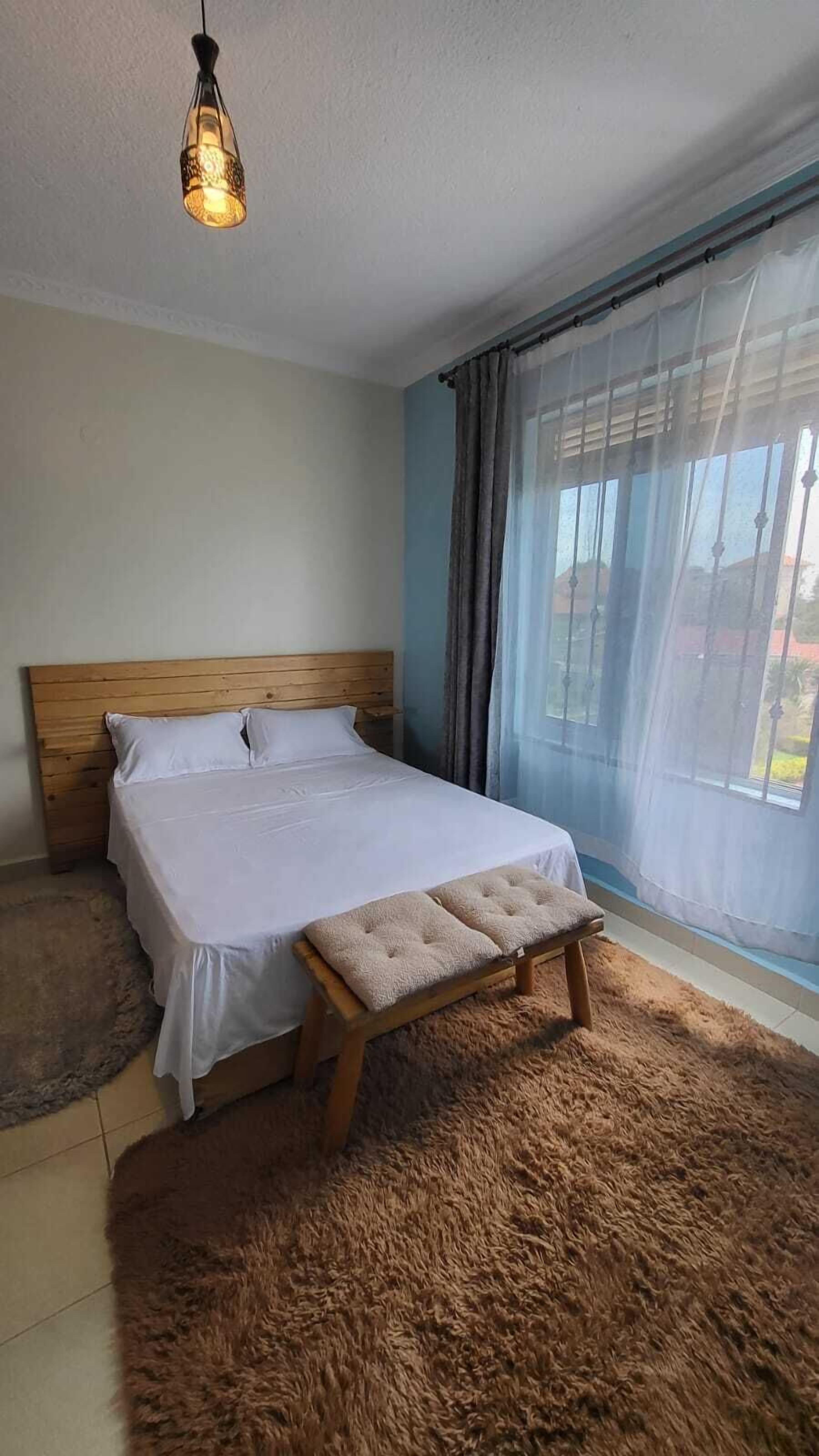 Ein Schlafzimmer mit einem hölzernen Bett, weißer Bettwäsche, einer Bank, einem flauschigen Teppich und einem Fenster mit durchsichtigen Vorhängen.