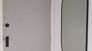 Une porte blanche avec une poignée noire et une petite plaque de verrouillage circulaire, un miroir ovale blanc fixé au mur, et un appareil de climatisation blanc placé au-dessus de la porte.