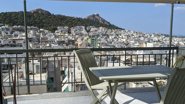 Un balcon avec une table et des chaises, donnant sur un paysage urbain avec des immeubles et une colline.