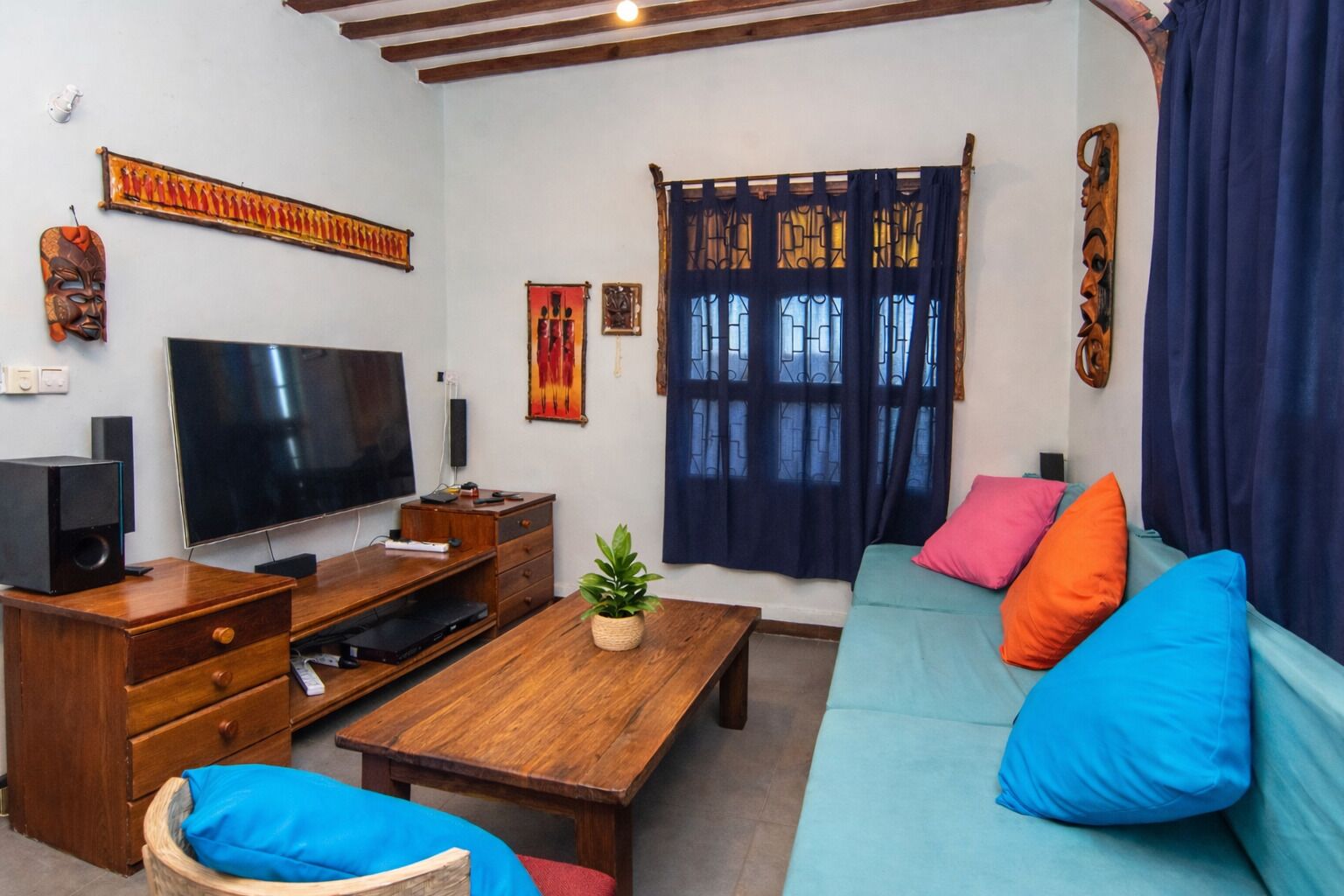 A living room with a TV, wooden furniture, a sofa with colorful cushions, and decorative items on the walls.