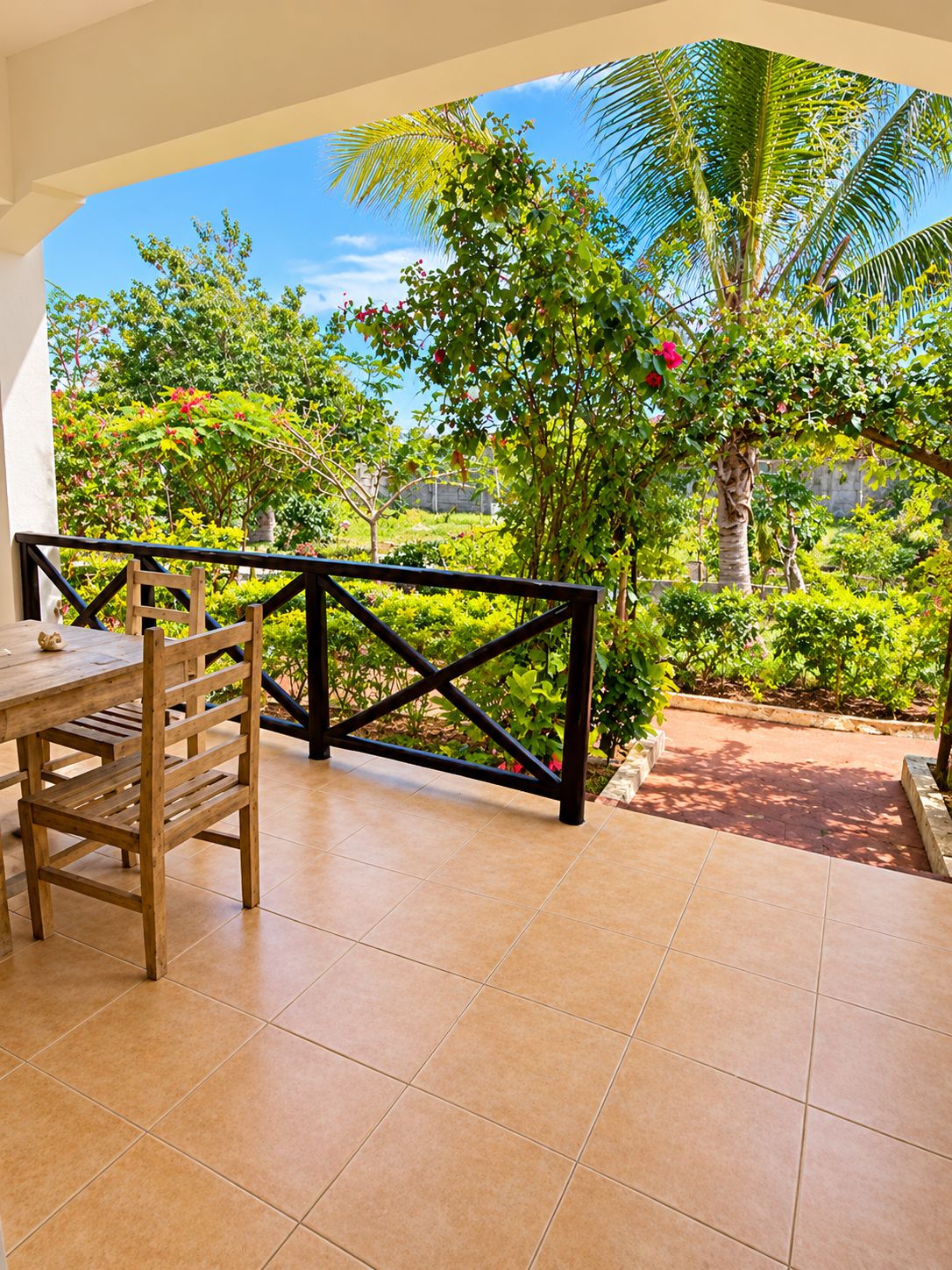 A covered patio with a wooden table and chairs, surrounded by lush greenery and a view of the sky.