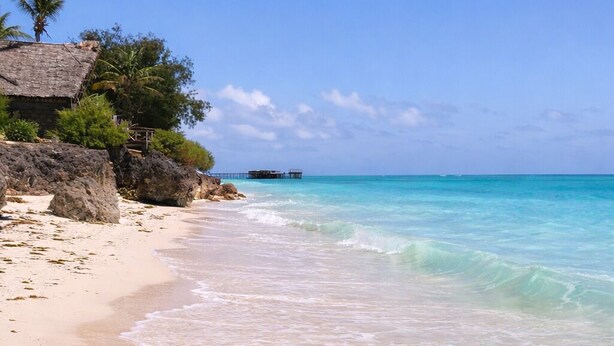 A beach with clear turquoise water, a sandy shore, and a structure with a thatched roof. - Majani Breeze Zanzibar (Nungwi)