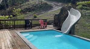 A pool with a slide, surrounded by a wooden deck and chairs.