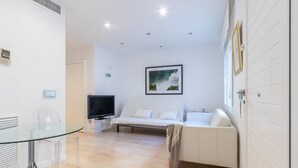 Une pièce moderne et bien éclairée, avec une table à manger en verre, un canapé blanc et un téléviseur posé sur un meuble.