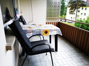 Ein Balkon mit Tisch, bedeckt mit einer Patchwork-Tischdecke, zwei schwarzen Stühlen und Blick auf eine Wohngegend.