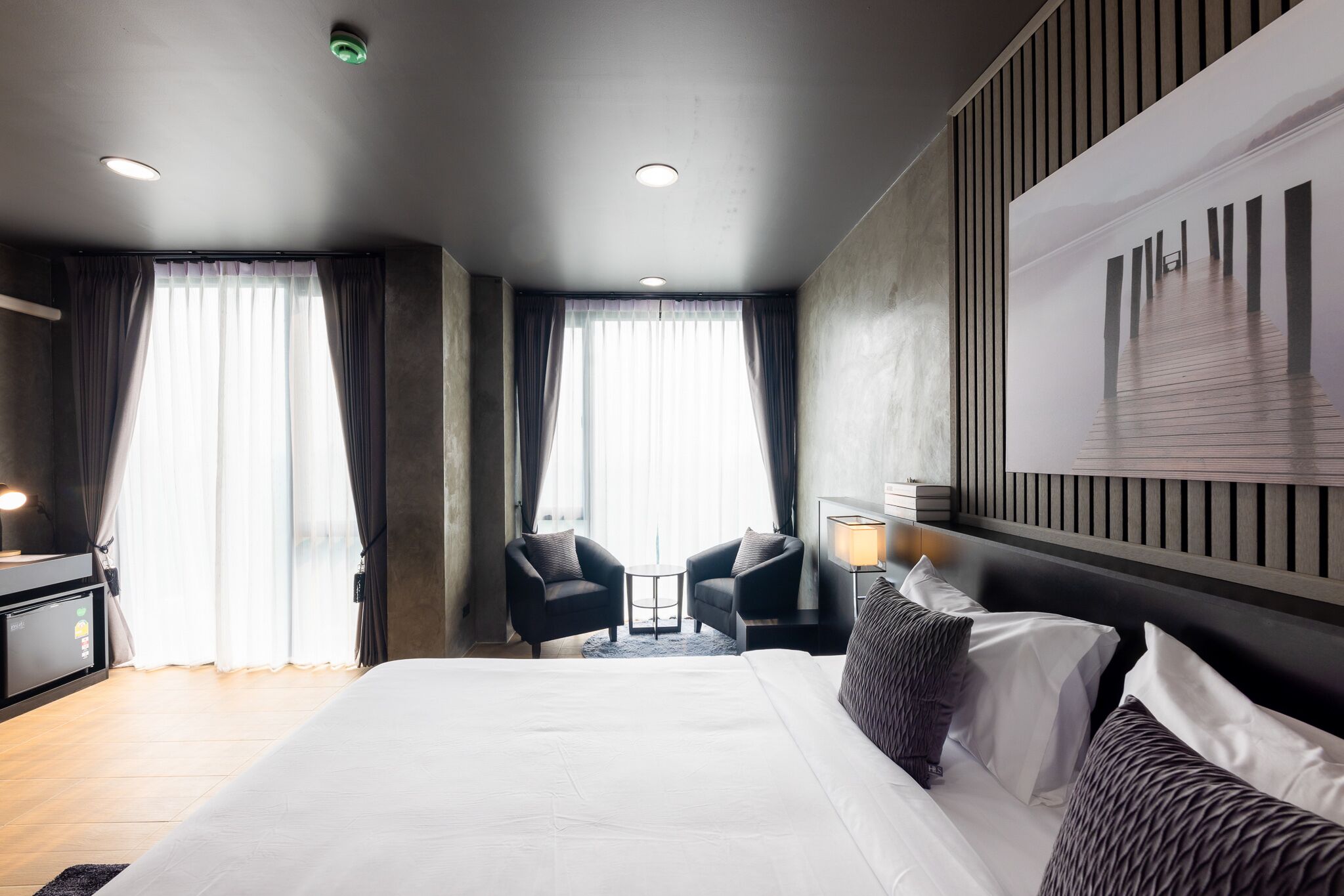 Quarto de hotel moderno com uma cama grande, duas poltronas, uma mesinha e vista para um edifício pela janela.