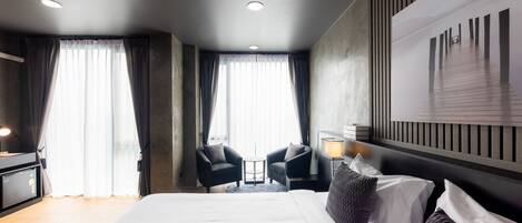 A modern hotel room with a large bed, two armchairs, a small table, and a view of a building through the window.
