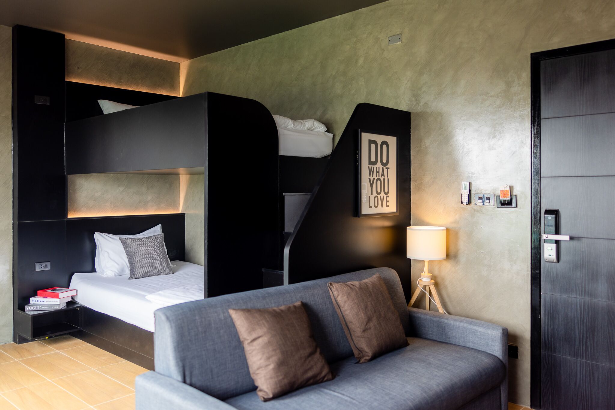 Una habitación de hotel moderna con una cama litera, un sofá, una mesita de noche con libros, una lámpara y un cartel motivacional.