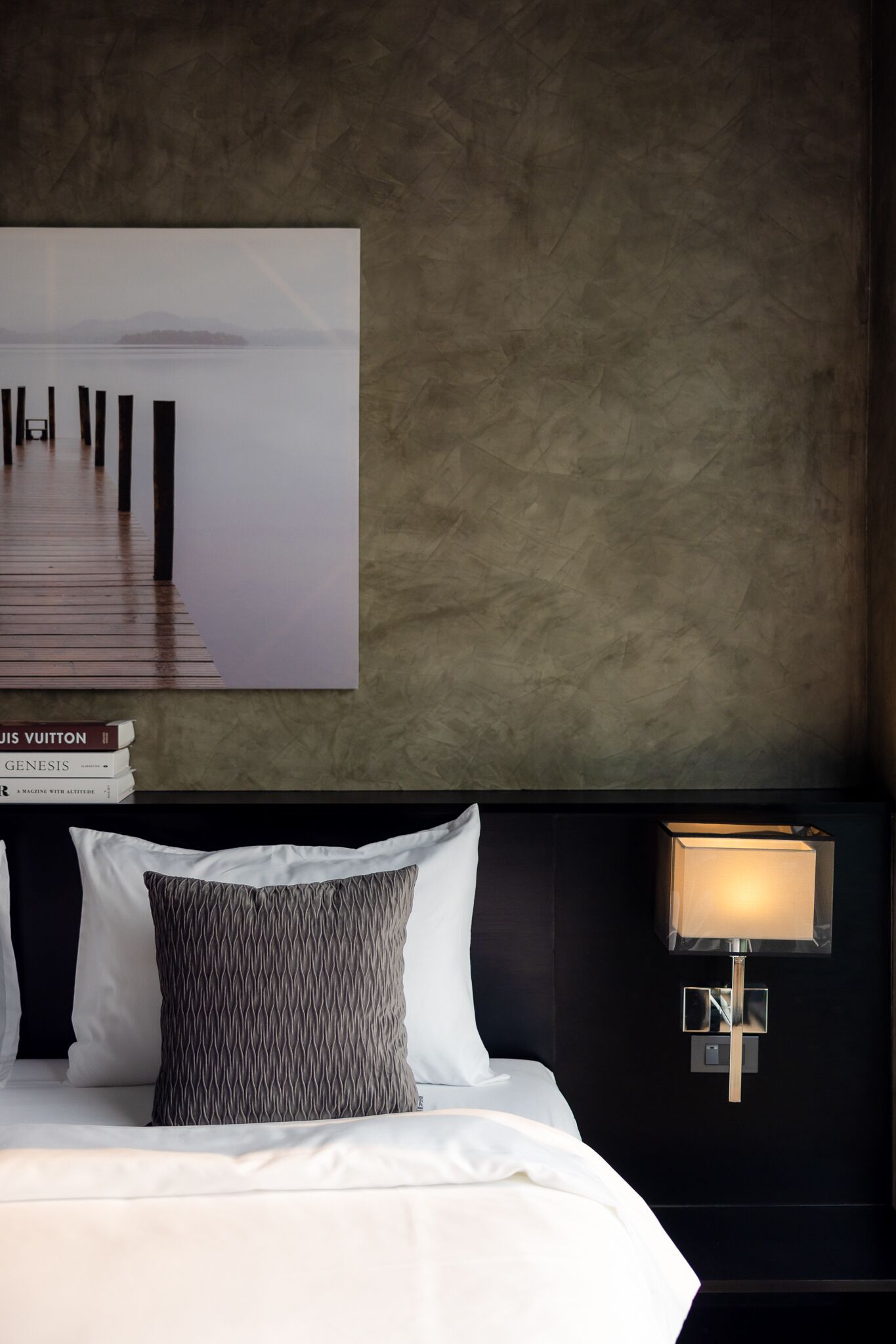 A hotel room with a large framed picture of a pier, a neatly made bed with white linens, and a modern wall-mounted lamp.