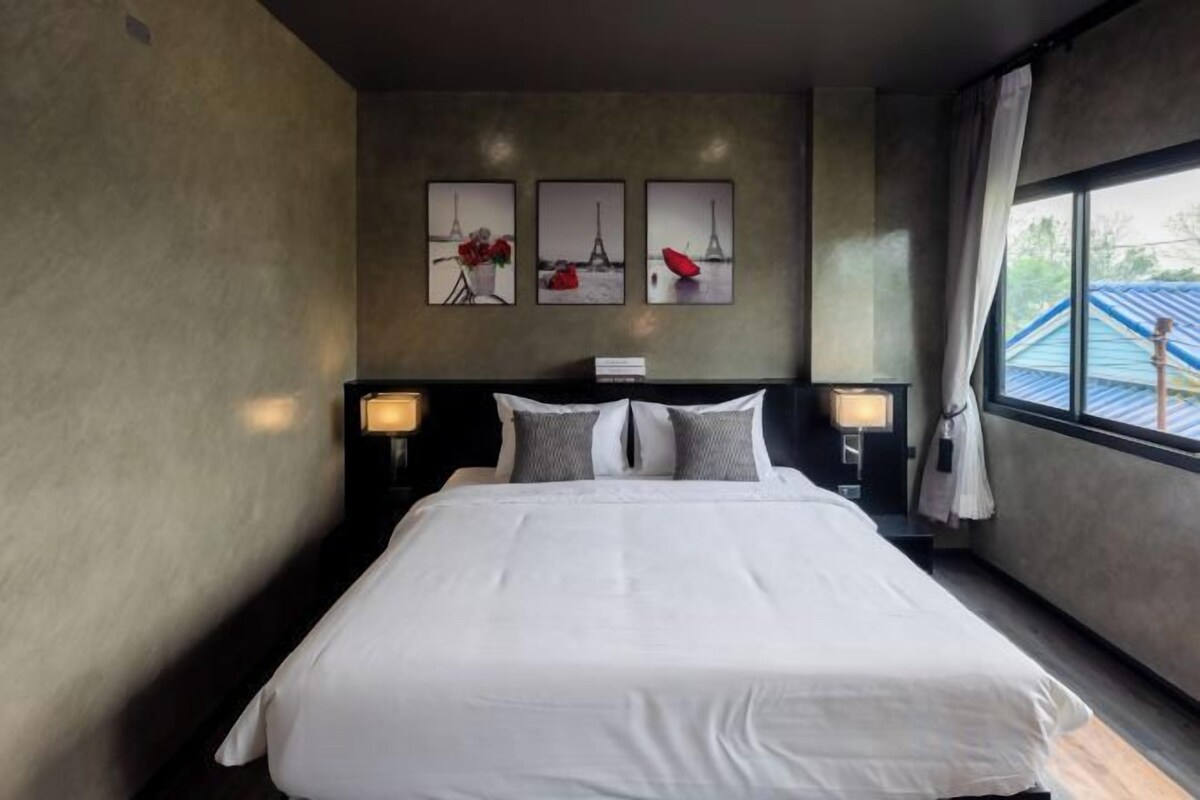 A hotel room with a large bed, two bedside tables with lamps, a window with curtains, and three wall-mounted pictures.