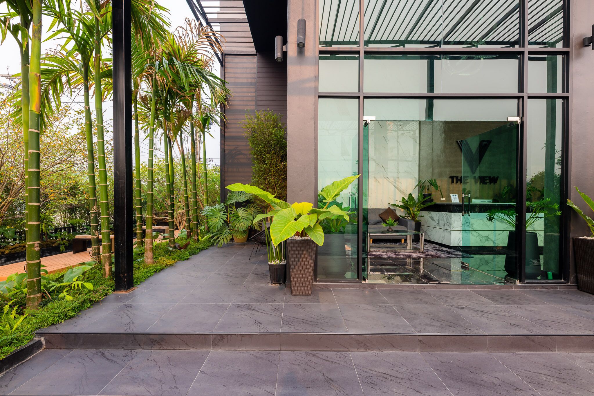 A modern building with a glass entrance, surrounded by a well-maintained garden with tall bamboo plants and large potted plants.