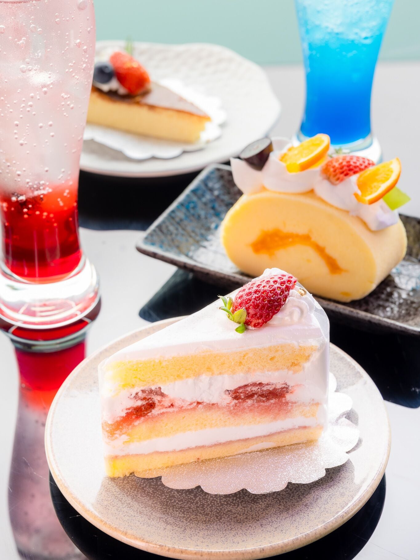 A slice of layered cake with strawberries on a plate, a glass of red beverage, and a blue drink in the background.