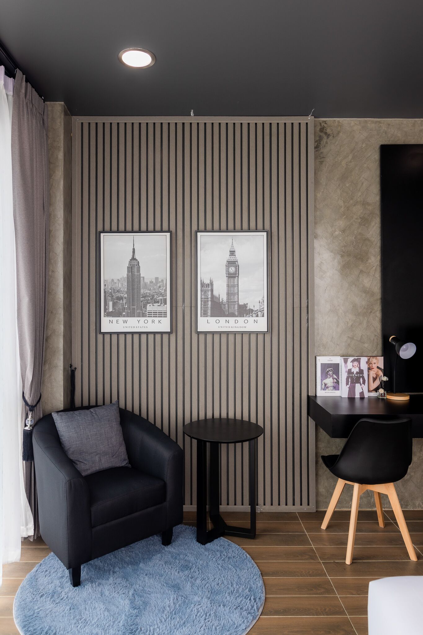 A modern room with a black leather sofa, a small round table, and a black desk with a chair. Two framed cityscape pictures adorn the wall, and a blue rug is on the floor.