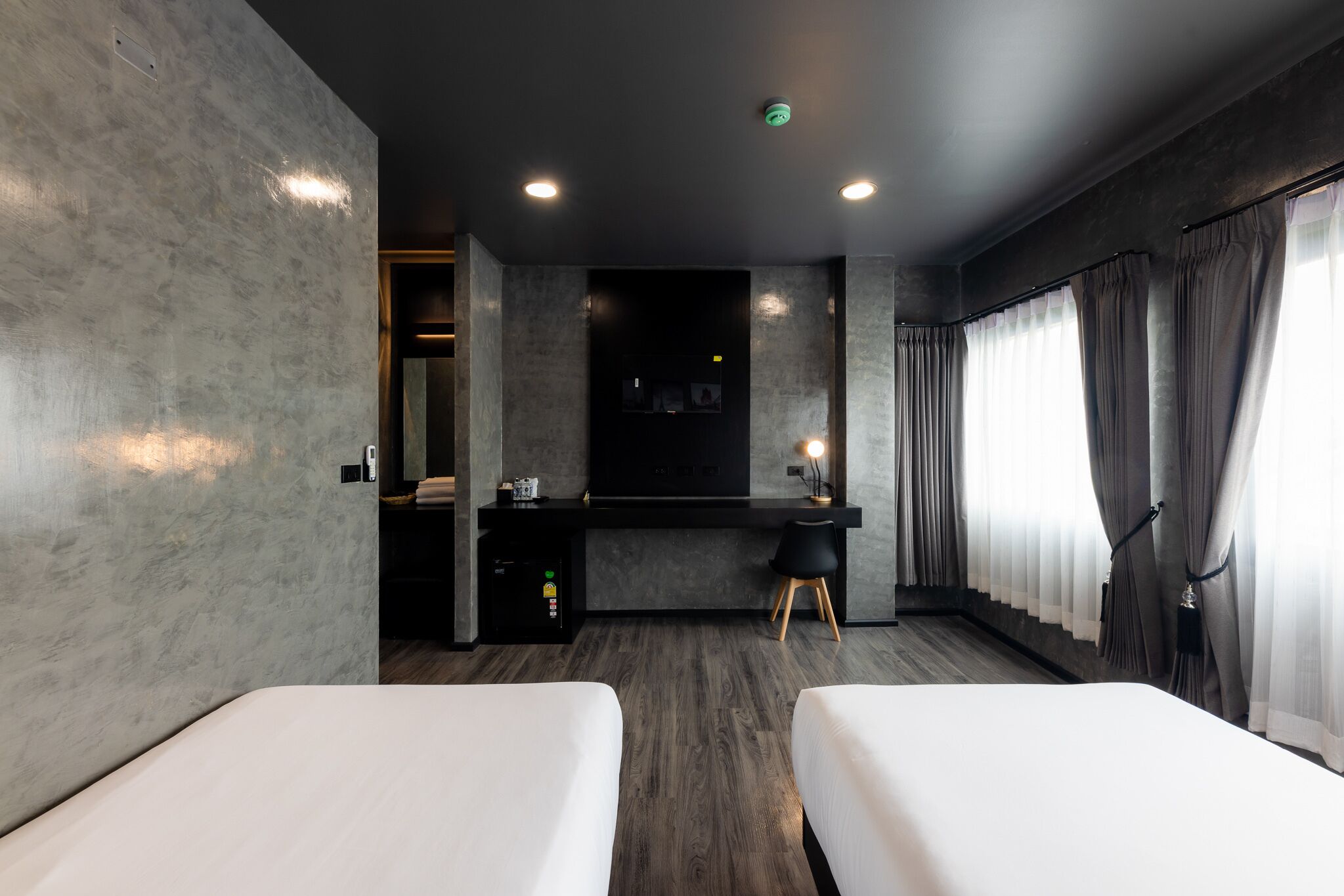 Moderni hotellihuone, jossa on kaksi erillistä sänkyä, työpöytä tuolilla, pieni keittokomero ja suuri ikkuna, jossa on verhot.