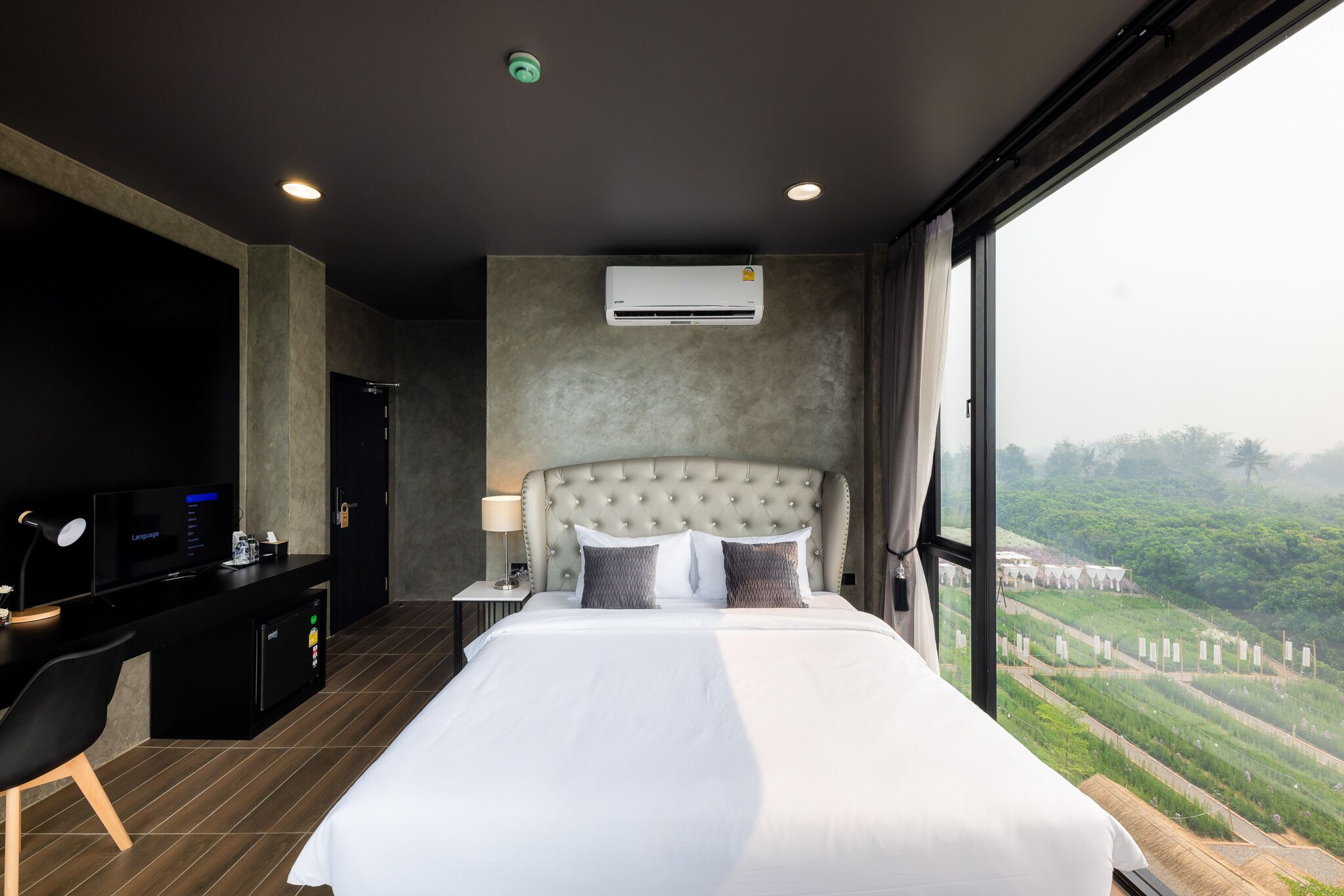 A modern hotel room with a large bed, a desk with a computer, and a view of a green landscape through a large window.