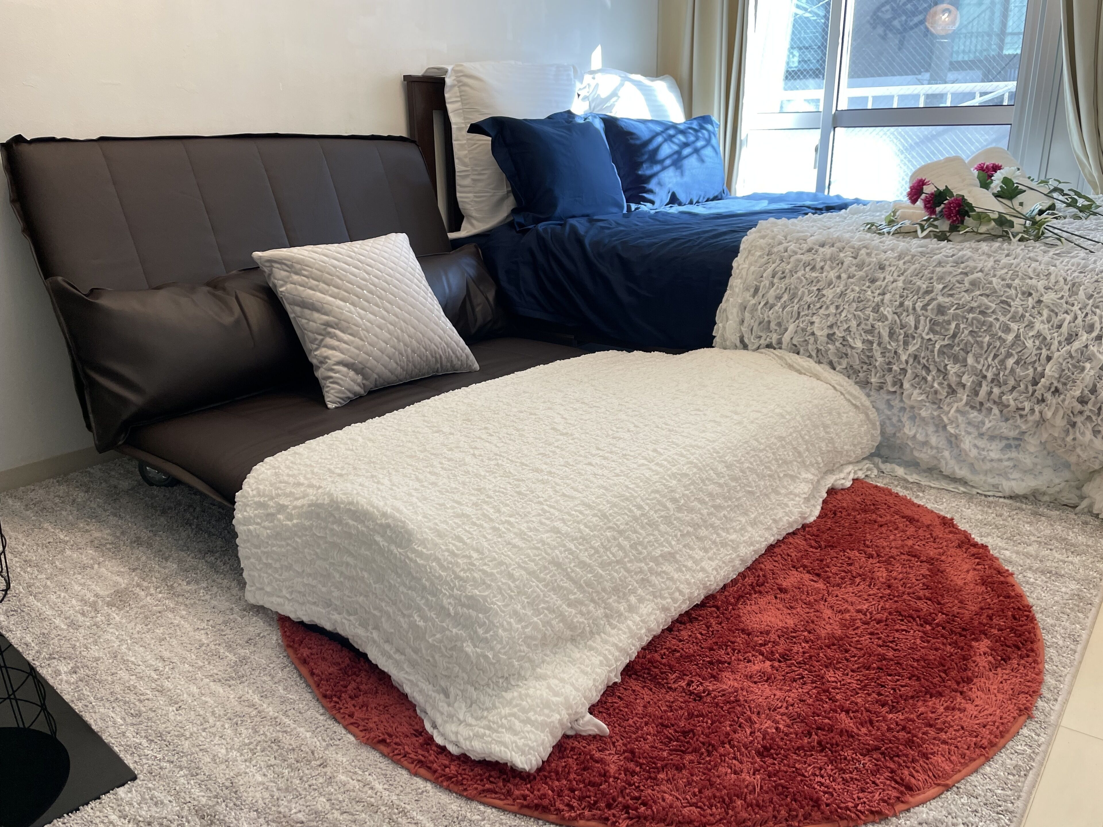 A bedroom with a brown sofa, a white blanket, a red rug, and a window with a view of the outside.