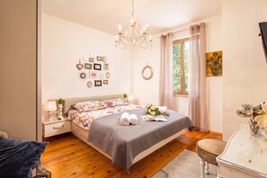Quarto com cama, mesas de cabeceira, lustre, janela com cortinas e decorações nas paredes.