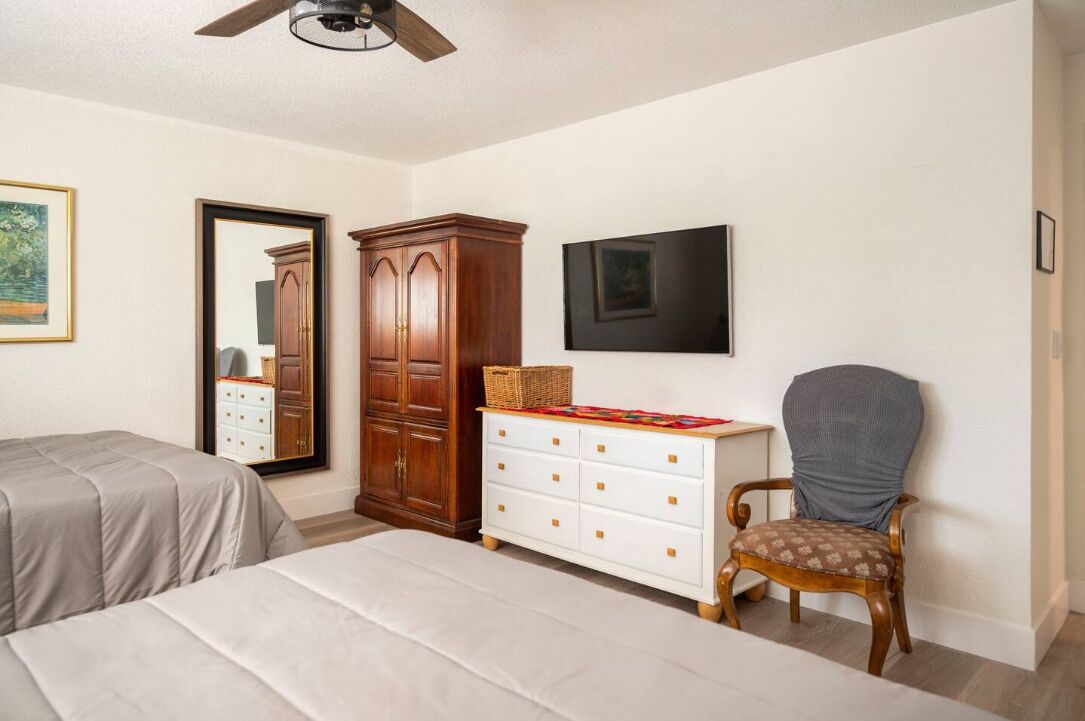 A bedroom with two beds, a wooden wardrobe, a white dresser, a chair, a ceiling fan, and a framed picture on the wall.