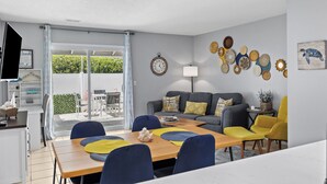 Ein modernes Wohnzimmer mit Essbereich, ein graues Sofa mit gelben Kissen, ein Holztisch und ein Fernseher auf einem Schrank.