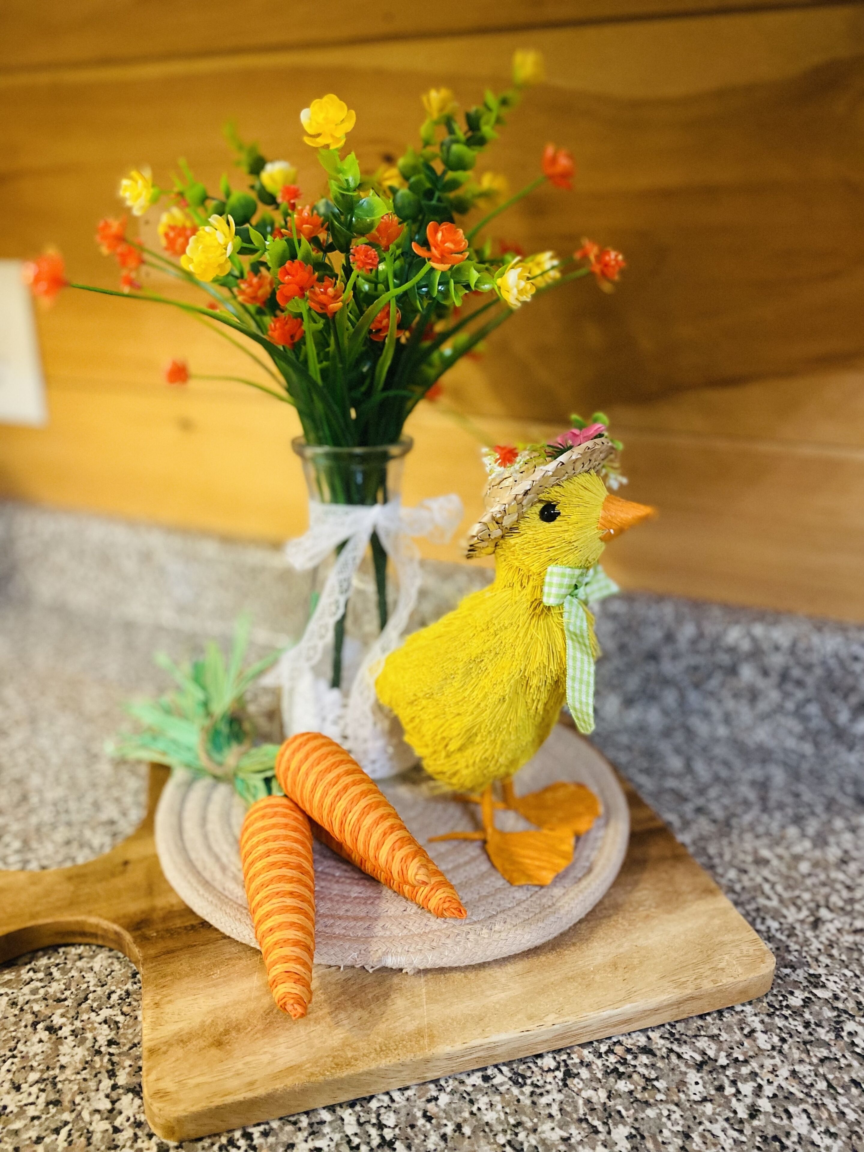 A decorative setup with a yellow chick figure, a vase of flowers, and carrots on a wooden board.